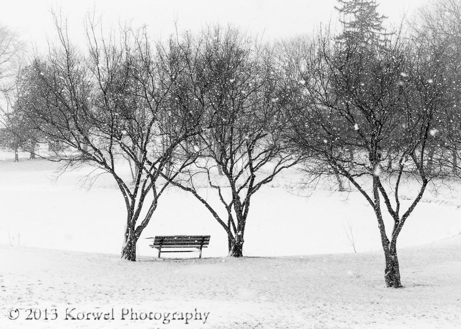 Snowing in tiny park with a bench